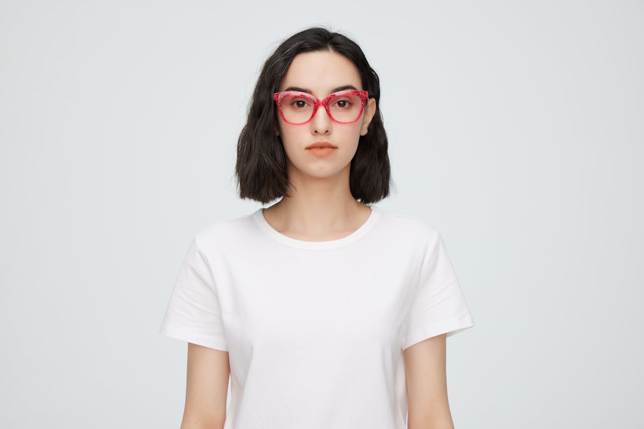 Portrait of a young woman wearing red eyeglasses and a white shirt against a plain background.